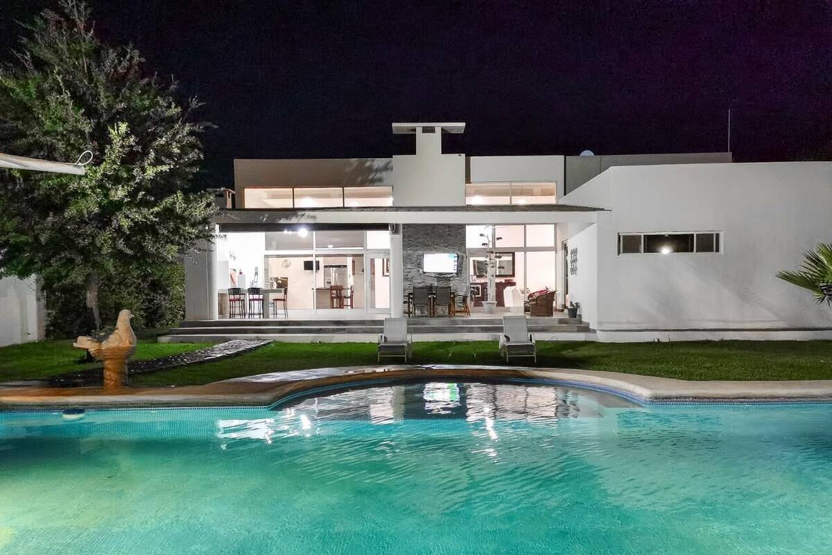 A modern house with a swimming pool, a dining area, and a well-lit exterior at night.