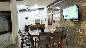 A dining area with a large table and chairs, a TV mounted on the wall, and a kitchen in the background.