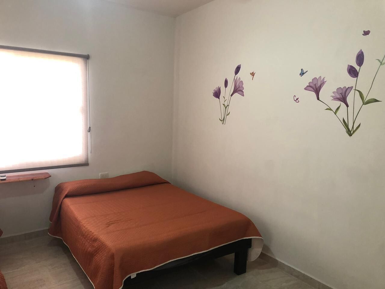 A small, single-bed room with a window, a bedside table, and a wall-mounted shelf.