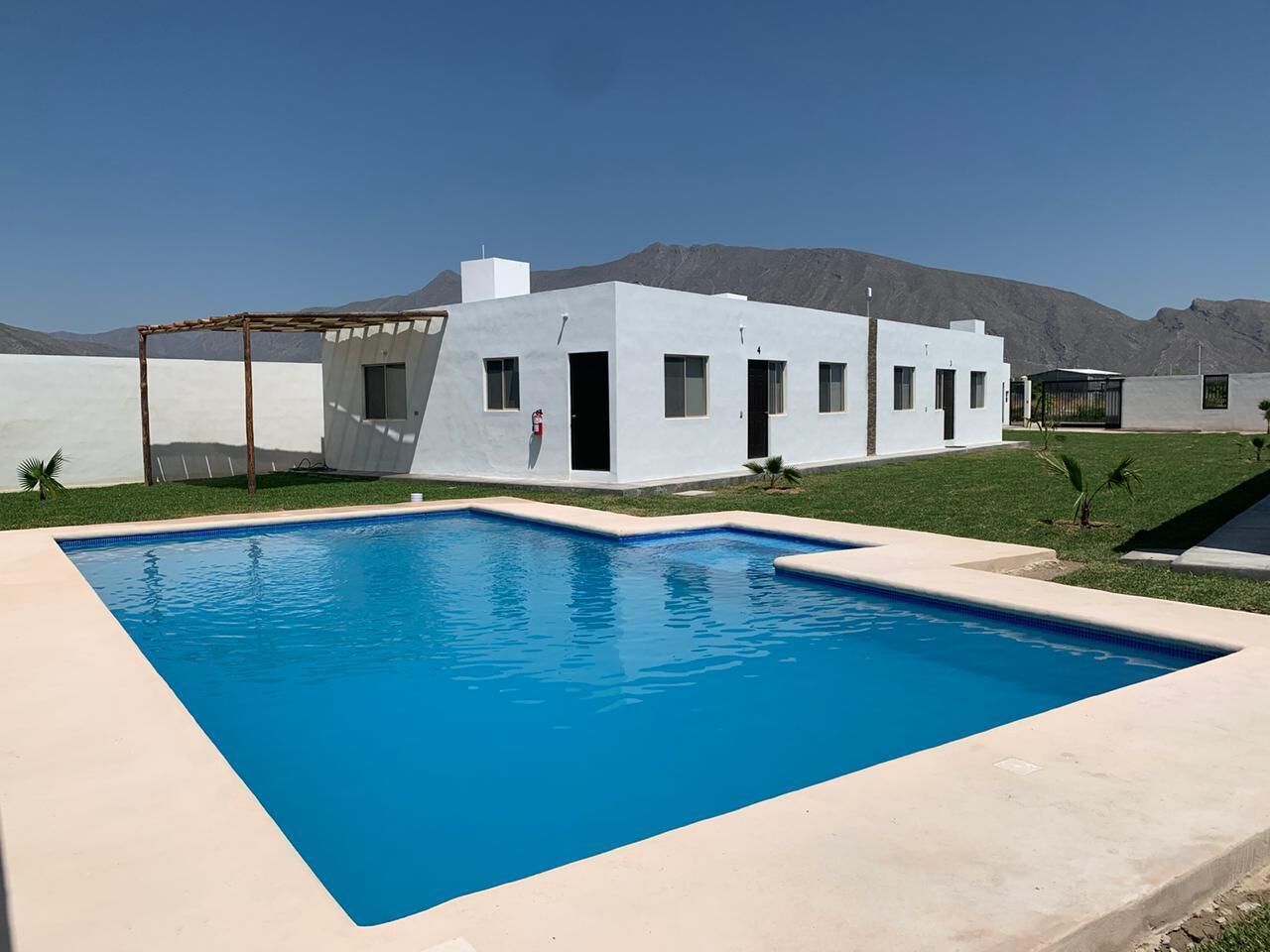 Un área de piscina moderna con una piscina rectangular, un edificio blanco con varias ventanas y un paisaje montañoso de fondo.