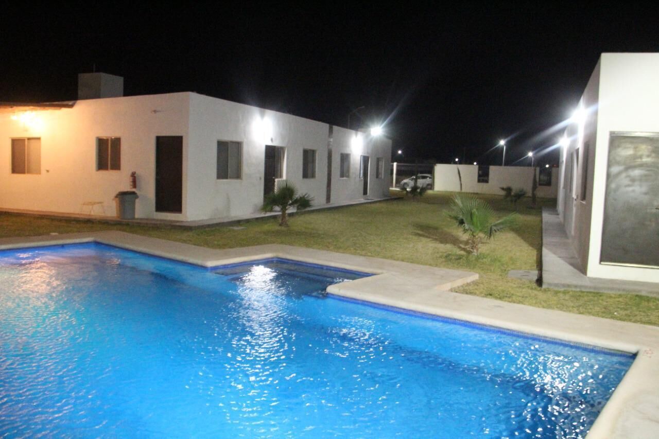 Una piscina con un edificio blanco al fondo, un pequeño césped y un coche aparcado cerca.