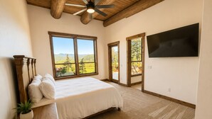 Un dormitorio con una cama grande, cabecera de madera, ventilador de techo, televisor en la pared y ventanas amplias con vistas al exterior.