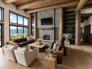 A spacious living room with a stone fireplace, large windows, and a high ceiling with exposed beams.