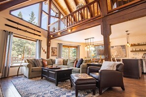 A spacious living room with a high ceiling, large windows, and a kitchen area visible through an open door. - Family A-Frame w/ Hot Tub & Game Loft near King’s Beach & Northstar (Tahoe Vista)