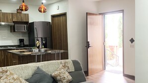 Une cuisine moderne équipée d’un four à micro-ondes encastré, d’une machine à café et d’un réfrigérateur. Le salon comprend un canapé gris agrémenté de coussins à motifs, ainsi qu’une porte-fenêtre coulissante donnant sur un espace extérieur.