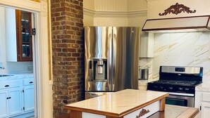 A kitchen with a central island, a brick accent wall, stainless steel appliances, and wooden bar stools. - The Historic Myrna Loy House, Downtown Hot Springs (Hot Springs)