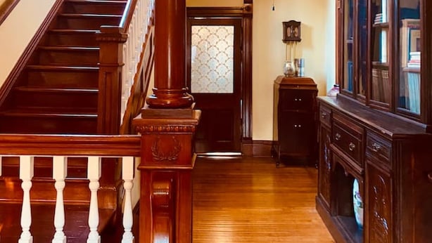 A grand staircase with wooden balusters, a ceiling fan, and a view into a room with a large wooden cabinet and a door with patterned glass. - The Historic Myrna Loy House, Downtown Hot Springs (Hot Springs)