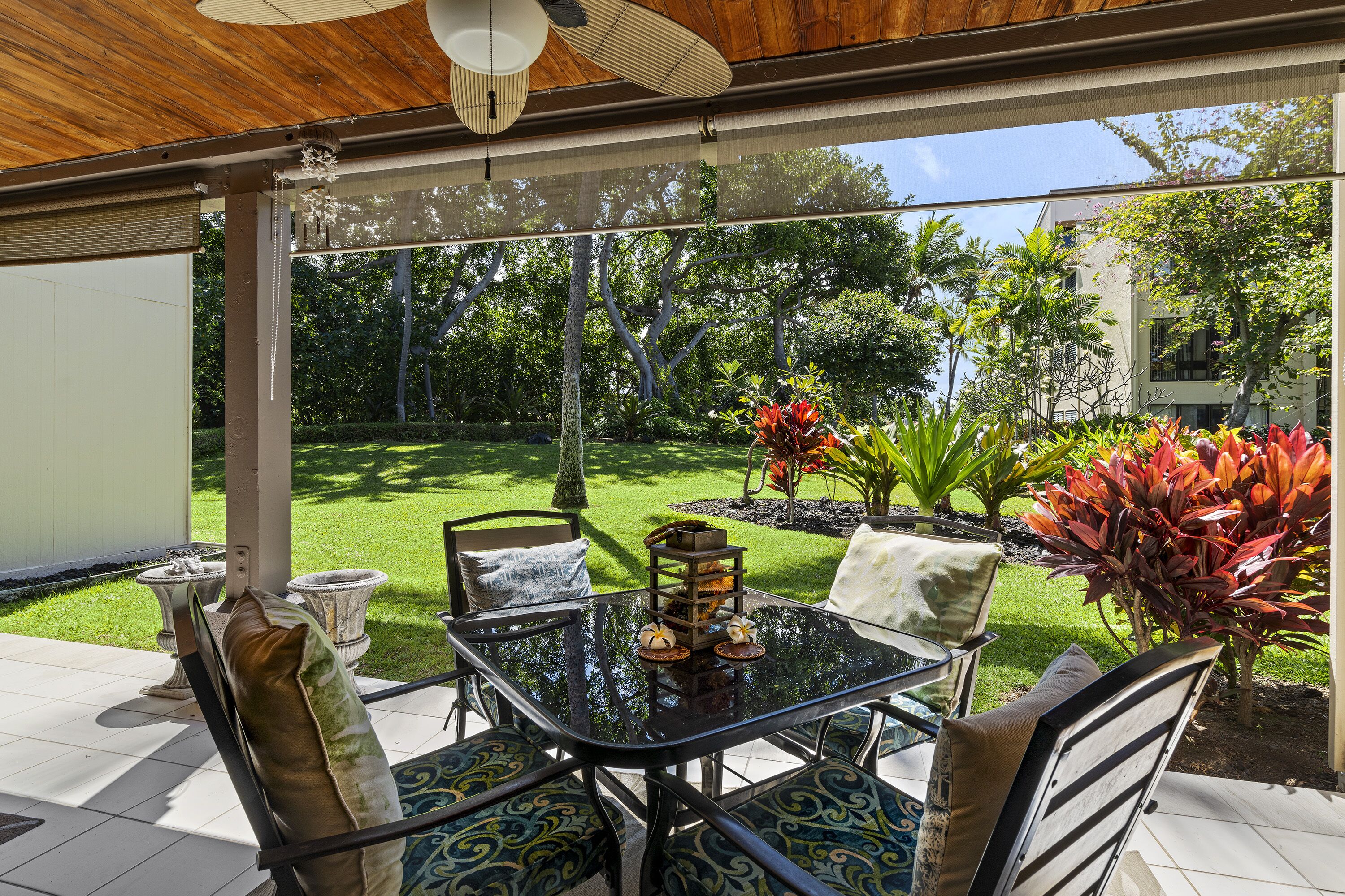 An outdoor patio with a glass table and chairs, surrounded by lush greenery and a view of a residential area.