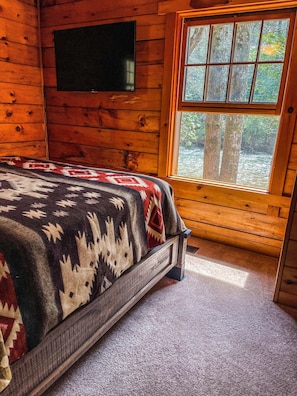 Habitación de cabaña de madera con una cama, un televisor montado en la pared y una ventana con vistas a árboles y agua.