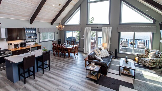 A spacious living area with a large dining table, a sofa, and a coffee table. There is a kitchen area with a stove and cabinets. The room has large windows and a view of the outdoors. - Cozy Lakefront! Lac La Nonne, Alberta (Barrhead County No.11)