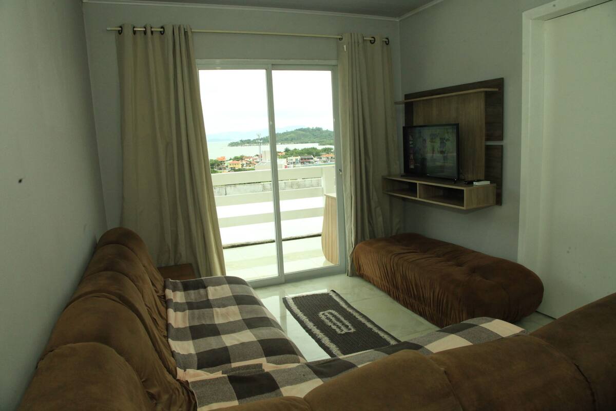A living room with a sofa, a television on a wooden shelf, and a view of a balcony with a railing.