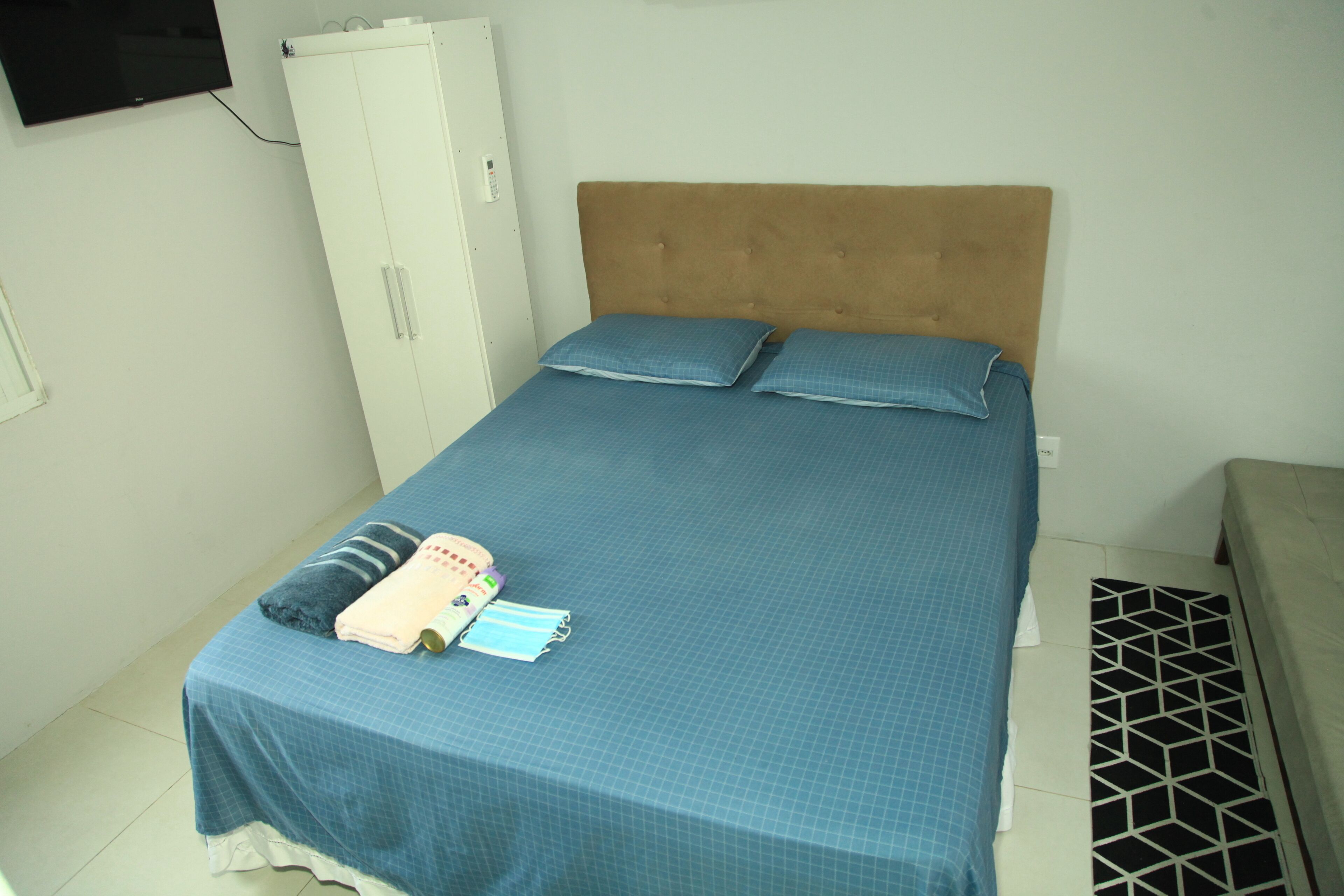 Una cama individual con colcha y almohadas azules, un armario blanco y un televisor montado en la pared.