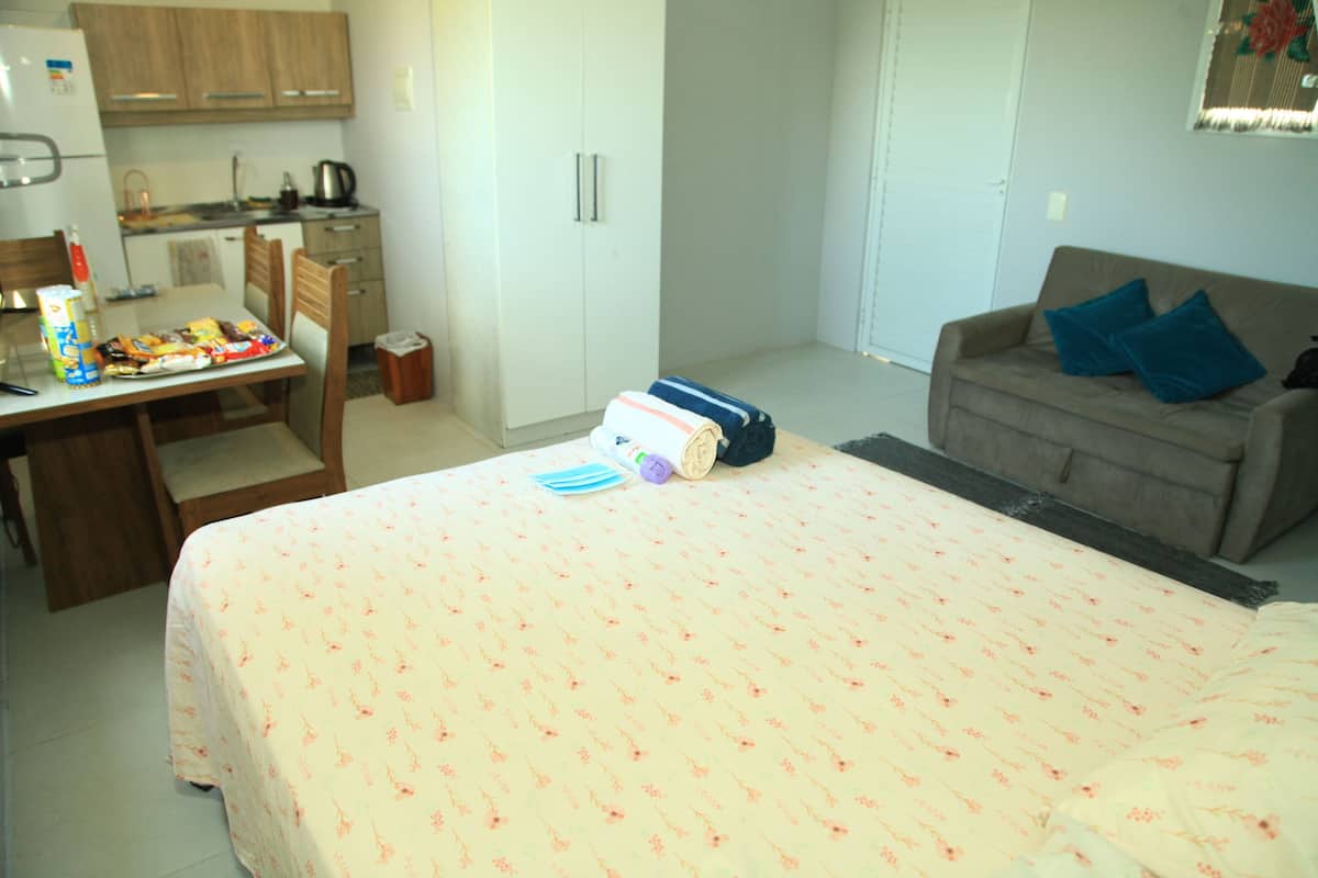 A bedroom with a bed, a sofa, a desk with a chair, a kitchen area, and a wardrobe.
