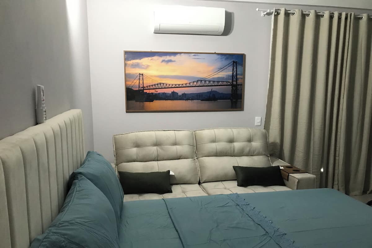 A bedroom with a bed, a nightstand, a painting of a bridge, and a window with curtains.