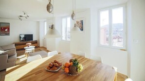 Un vaste espace de vie comprenant une table à manger en bois, dressée avec des fruits et des pâtisseries, un canapé et un téléviseur en arrière-plan.