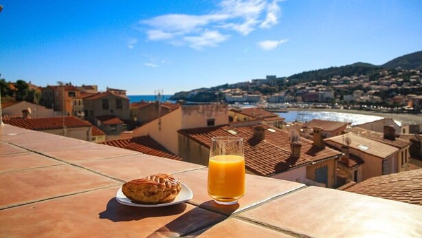 Une table avec une pâtisserie et un verre de jus d’orange, donnant sur une ville côtière aux toits de tuiles rouges et sous un ciel bleu dégagé.