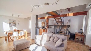 Un vaste espace de vie comprenant un canapé, un pouf et une table à manger. On aperçoit une cuisine au fond, et un escalier mène à l’étage supérieur.