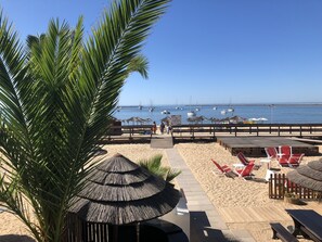 A beachside area with a wooden deck, beach chairs, umbrellas, and a view of the sea with boats.