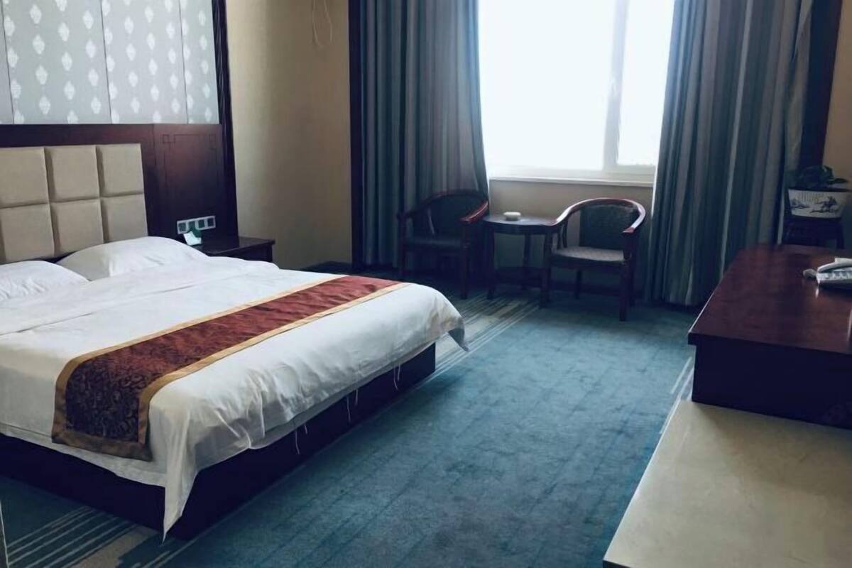 Ein Hotelzimmer mit einem Bett, einem Schreibtisch, zwei Stühlen, einem Vorhangfenster und einem an der Kopfteil befestigten Licht.