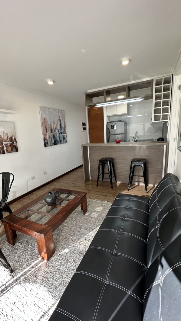 A modern living room with a black leather sofa, a wooden coffee table, and a glass-top dining table. There are two black stools and a kitchen area with a white countertop and a sink.