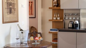 Un coin repas avec une table dressée pour deux personnes, une bouteille de champagne et un plateau de desserts. On y trouve des livres et des ustensiles de cuisine sur des étagères, ainsi qu’une machine à café sur le comptoir.