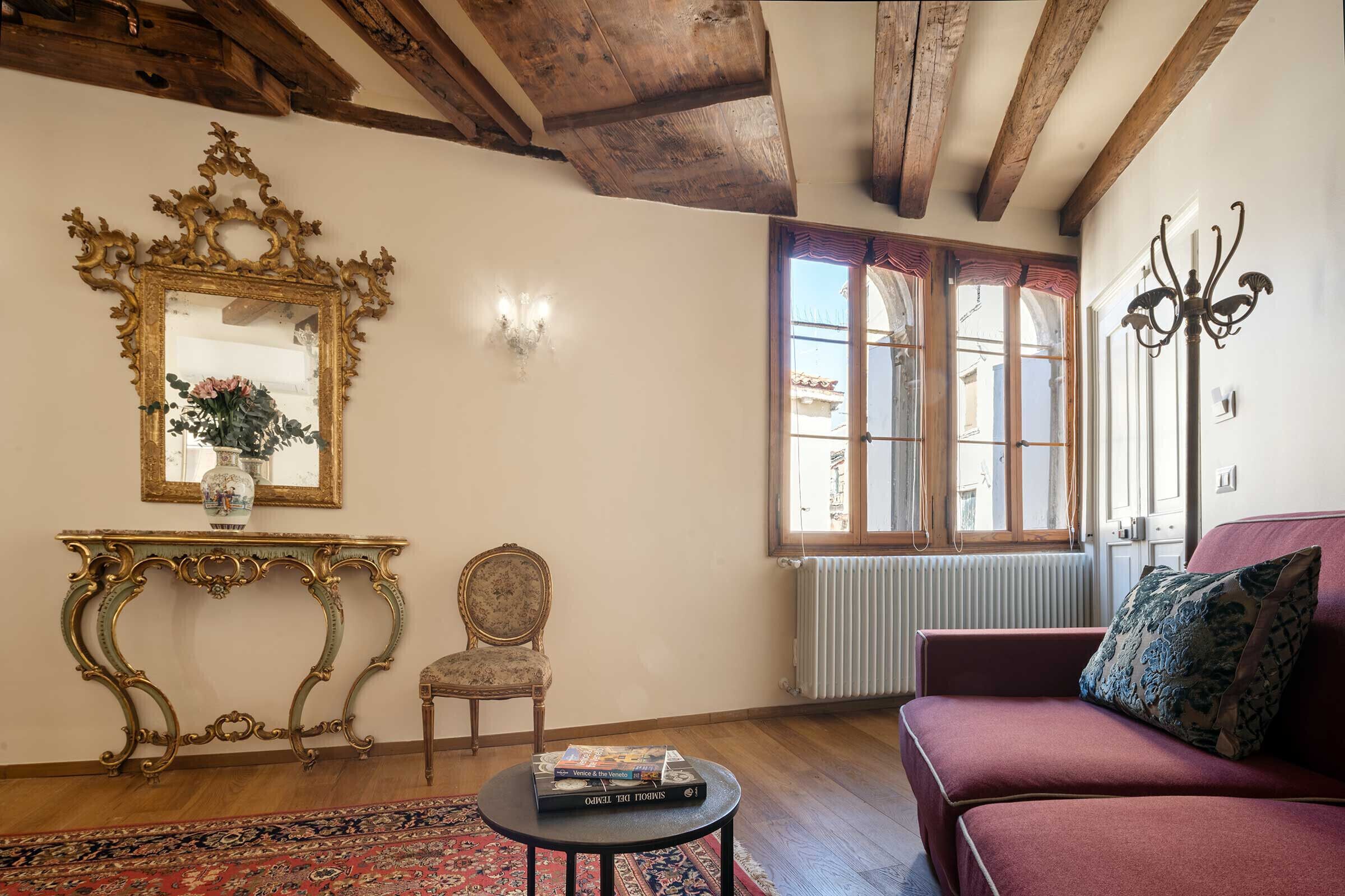 A room with a wooden ceiling, a large mirror, a sofa, a chair, and a small table with books.