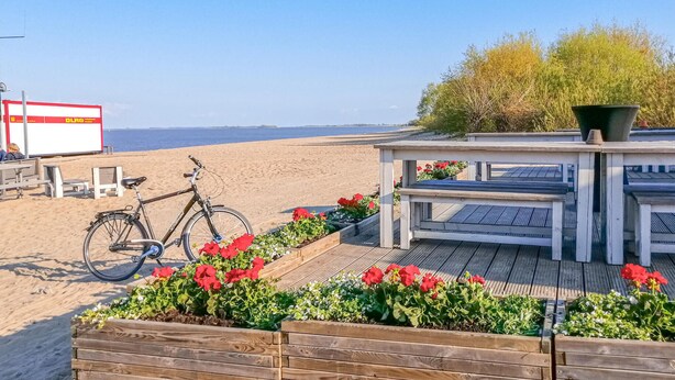 Terraza frente al mar con bancos de madera y una bicicleta, con vista a una playa de arena y al océano.
