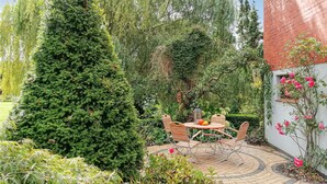 Un patio con una mesa redonda y sillas, rodeado de abundante vegetación y un alto árbol de coníferas.