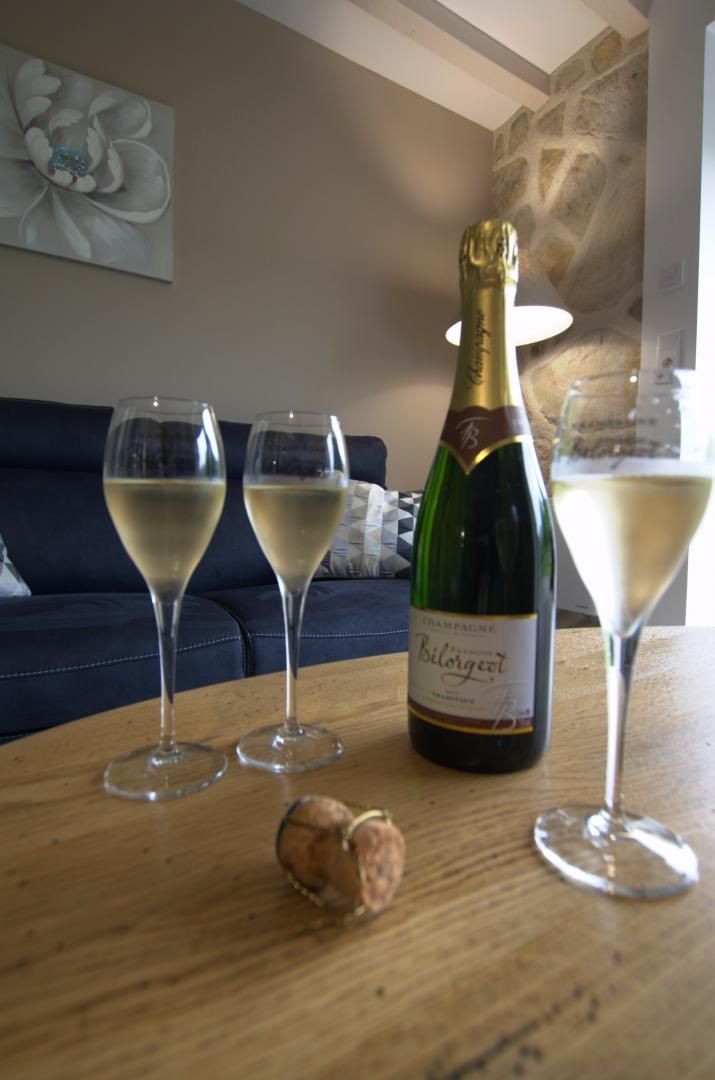 A bottle of champagne, two glasses of champagne, and a cork on a wooden table in a room with a blue couch and a stone wall.