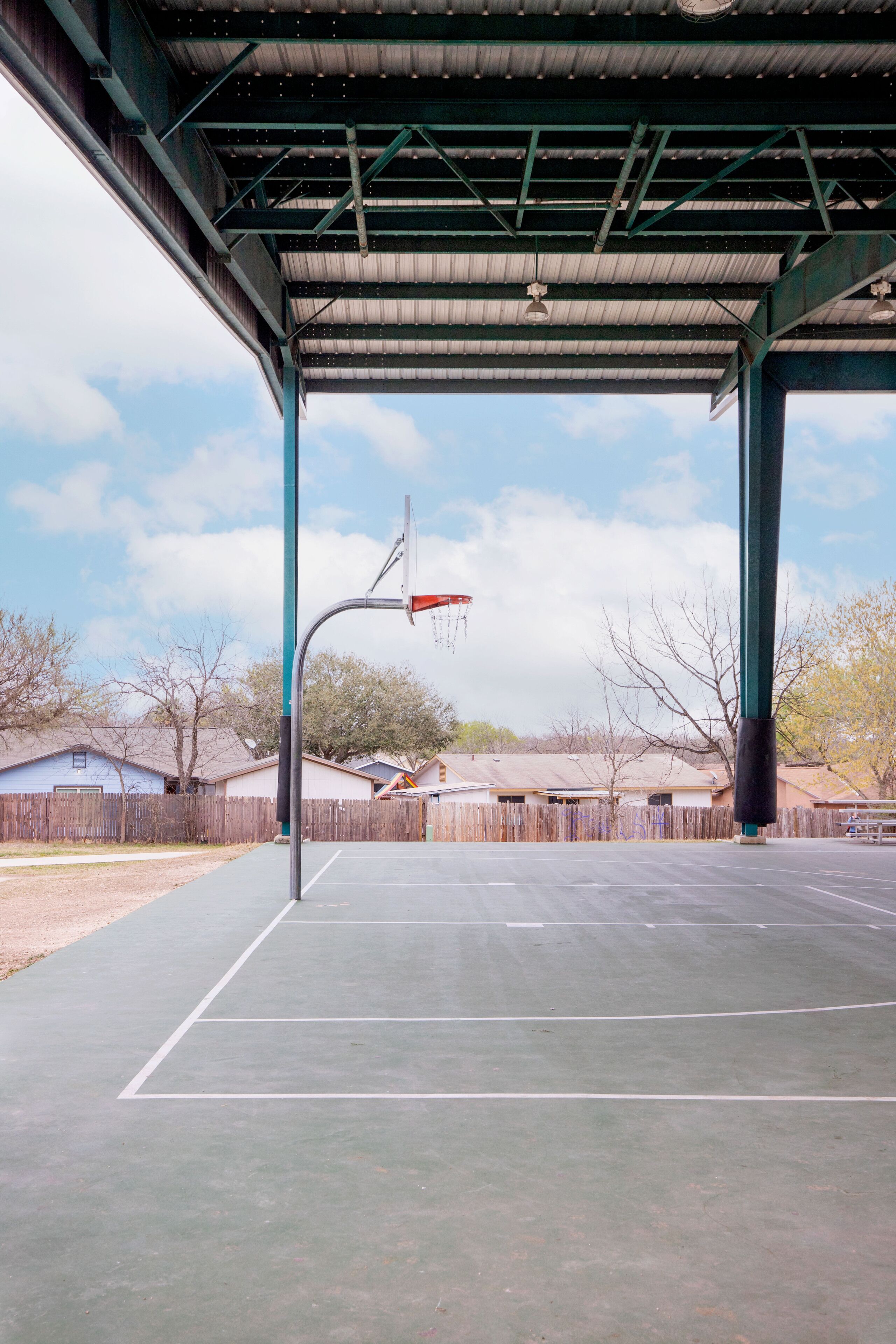 An outdoor basketball court with a metal roof structure, surrounded by a wooden fence and residential buildings in the background.
