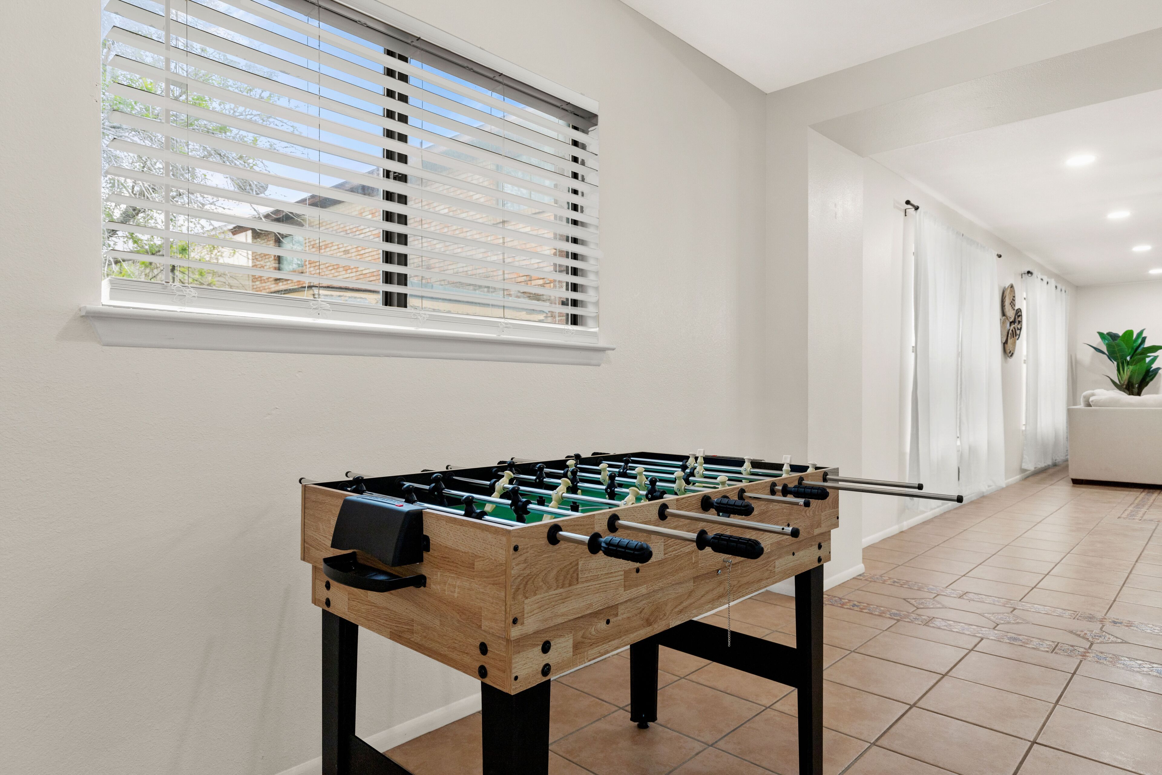 Uma mesa de pebolim em um ambiente com paredes brancas, uma janela com persianas e piso de azulejos.