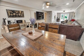 A dining area with a wooden table, chairs, and a vase with flowers. There is a sofa and a television in the background.