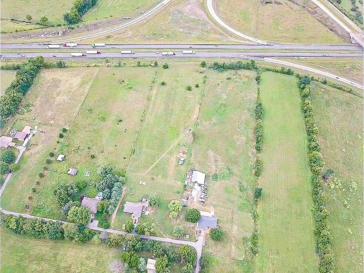 Vue aérienne d’une zone rurale avec un vaste champ ouvert, quelques bâtiments dispersés et une autoroute en arrière-plan.