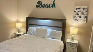 A neatly made bed with white bedding, two bedside tables with lamps, a wall-mounted sign reading "Beach", and a framed artwork of sea turtles. - The Dunes 508 - Beach View & Bay View-Indoor-Outdoor Pool (Gulf Shores)