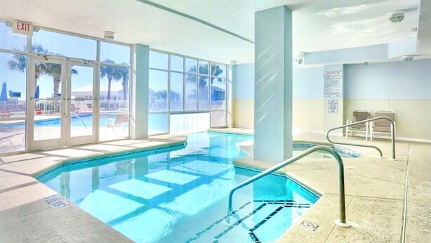 An indoor swimming pool with clear glass walls, a visible exit sign, and a poolside area with chairs. - The Dunes 508 - Beach View & Bay View-Indoor-Outdoor Pool (Gulf Shores)