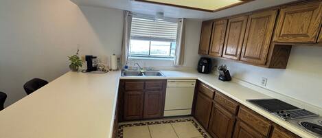 A compact kitchen with a dishwasher, a sink, wooden cabinets, a window with blinds, and a countertop with a coffee maker.