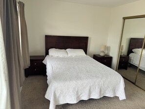 Una cama bien hecha con colcha blanca, cabecera de madera, mesita de noche con lámpara y un espejo grande en la pared.