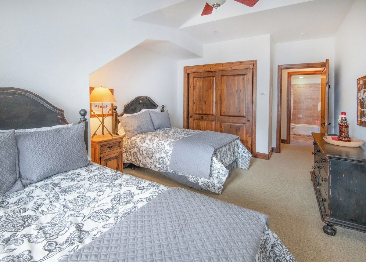 A bedroom with two beds, a nightstand with a lamp, a dresser, and a wooden door.