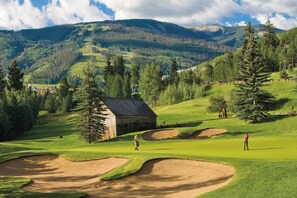 A golf course with bunkers, a wooden building, and a mountainous backdrop.