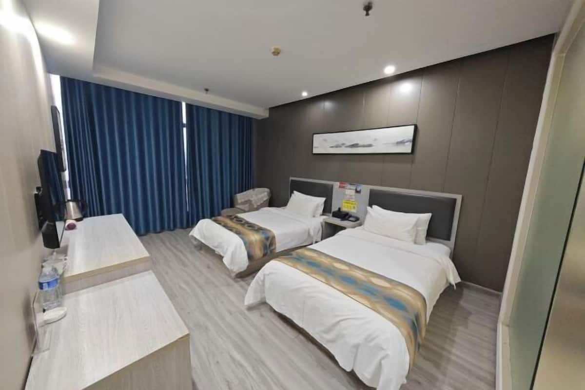 A hotel room with two beds, a desk, a flat-screen TV, and a large window with blue curtains.