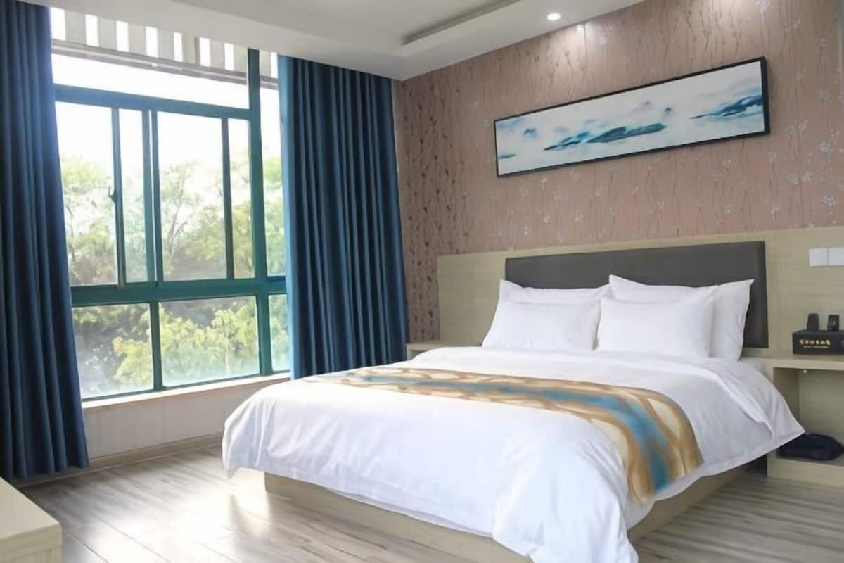 A hotel room with a large bed, a window with curtains, a headboard, and a painting on the wall.