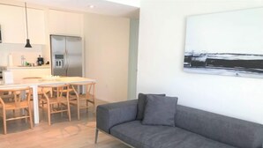 Uma sala de estar moderna com um sofá cinzento, mesa de jantar de madeira e cadeiras. Cozinha com frigorífico e uma luminária pendente visível ao fundo.