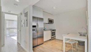Cozinha moderna com frigorífico, micro-ondas, forno e pia. Há uma mesa de jantar com cadeiras e uma janela com persianas.
