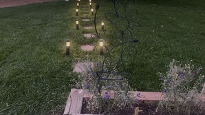 Een tuin met een metalen sculptuur, een houten plantenbak en een pad met verlichting.