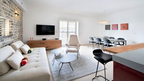 Una sala moderna con sofá, mesa de centro y sillones, un televisor de pantalla plana en la pared, y un comedor con mesa y sillas de madera.