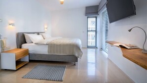 Un dormitorio moderno con cama, mesitas de noche, ventilador de techo, televisor montado en la pared y ventana con persianas.