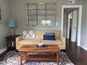 A living room with a beige sofa, a blue pillow, a wooden coffee table, a lamp, and a large window with a distressed frame.