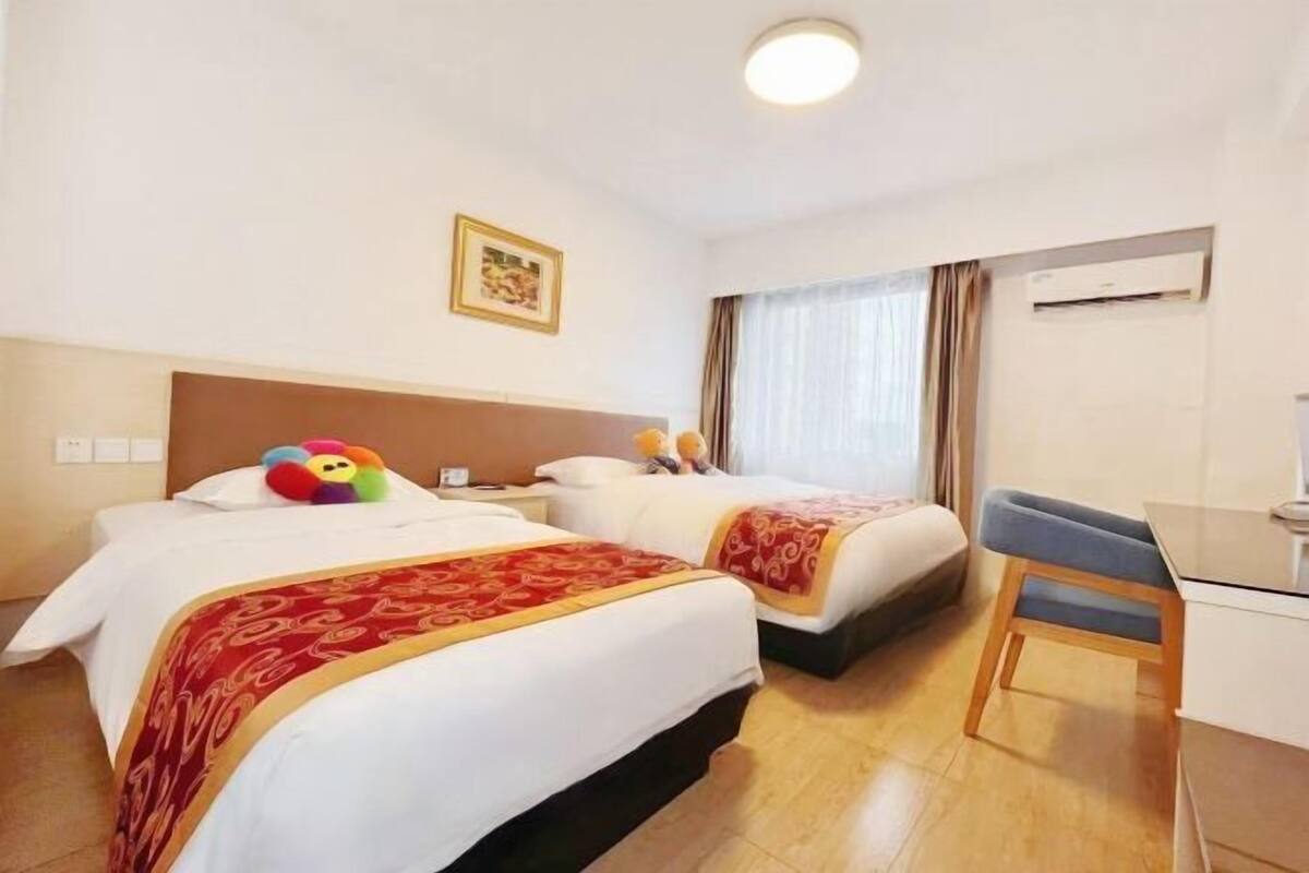 Hotellihuoneessa on kaksi sänkyä, työpöytä ja tuoli. Huoneessa on ikkuna, jossa on verhot, ilmastointilaite ja seinällä kehystetty taulu.