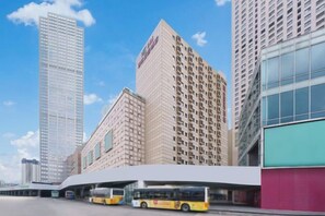 Un edificio moderno con fachada curva, una estación de autobuses en primer plano y edificios altos al fondo.