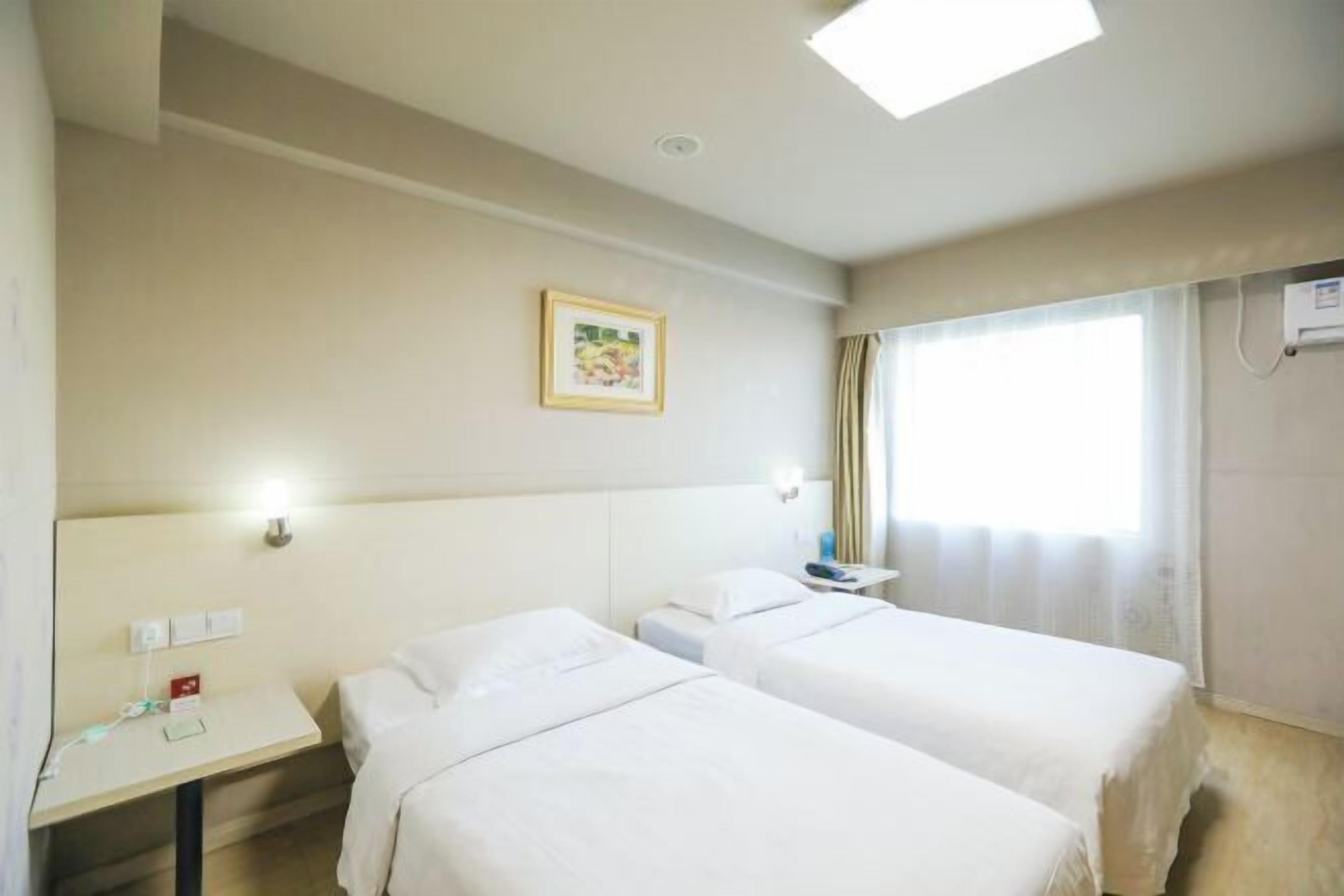 A hotel room with two beds, a bedside table, a small desk, a window with curtains, and a ceiling light.